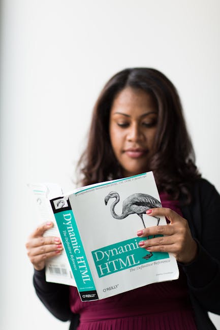A woman with medium skin tone and long, dark brown hair stands indoors against a plain white wall, holding an open book titled 'Dynamic HTML' with one hand on the left page and the other on the right 